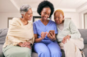 Smiling caregiver in blue scrubs sitting on a couch with two senior women, showing them something on a smartphone. A moment of connection and engagement in a warm home care setting.