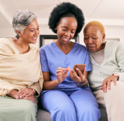 Smiling caregiver in blue scrubs sitting on a couch with two senior women, showing them something on a smartphone. A moment of connection and engagement in a warm home care setting.