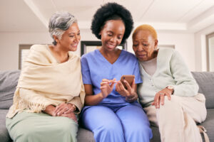 Smiling caregiver in blue scrubs sitting on a couch with two senior women, showing them something on a smartphone. A moment of connection and engagement in a warm home care setting.